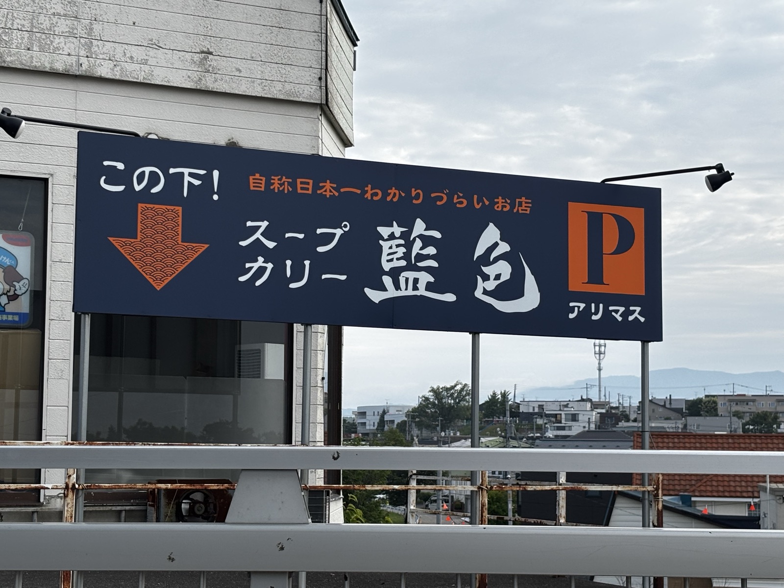 藍色の看板。「この下!自称日本一わかりづらいお店」と書いてある